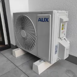 Zewnętrzna jednostka klimatyzatora marki AUX, zamontowana na białych wspornikach na szarym balkonie, widoczna ściana budynku z szarą fakturą.