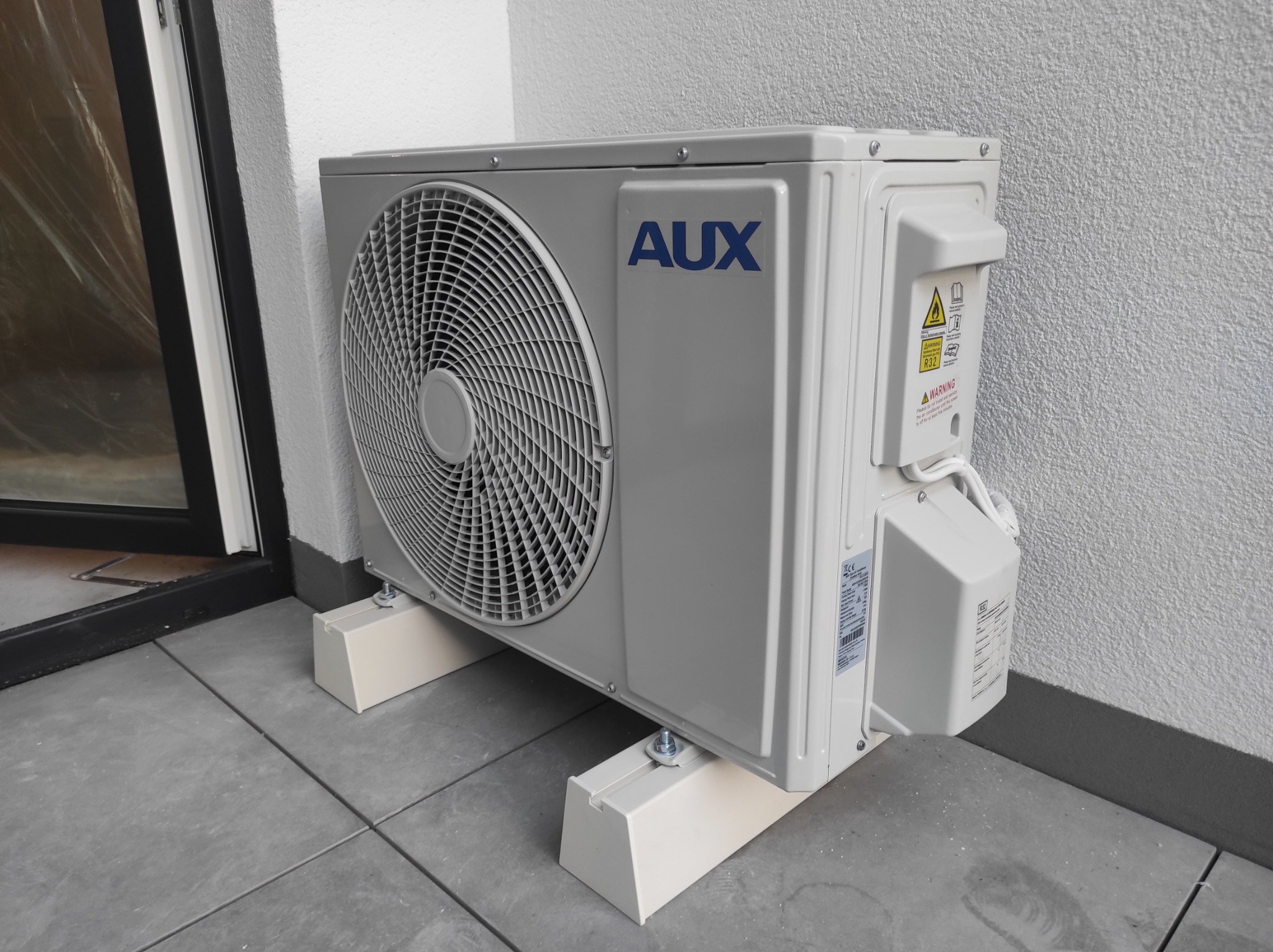Zewnętrzna jednostka klimatyzatora marki AUX, zamontowana na białych wspornikach na szarym balkonie, widoczna ściana budynku z szarą fakturą.