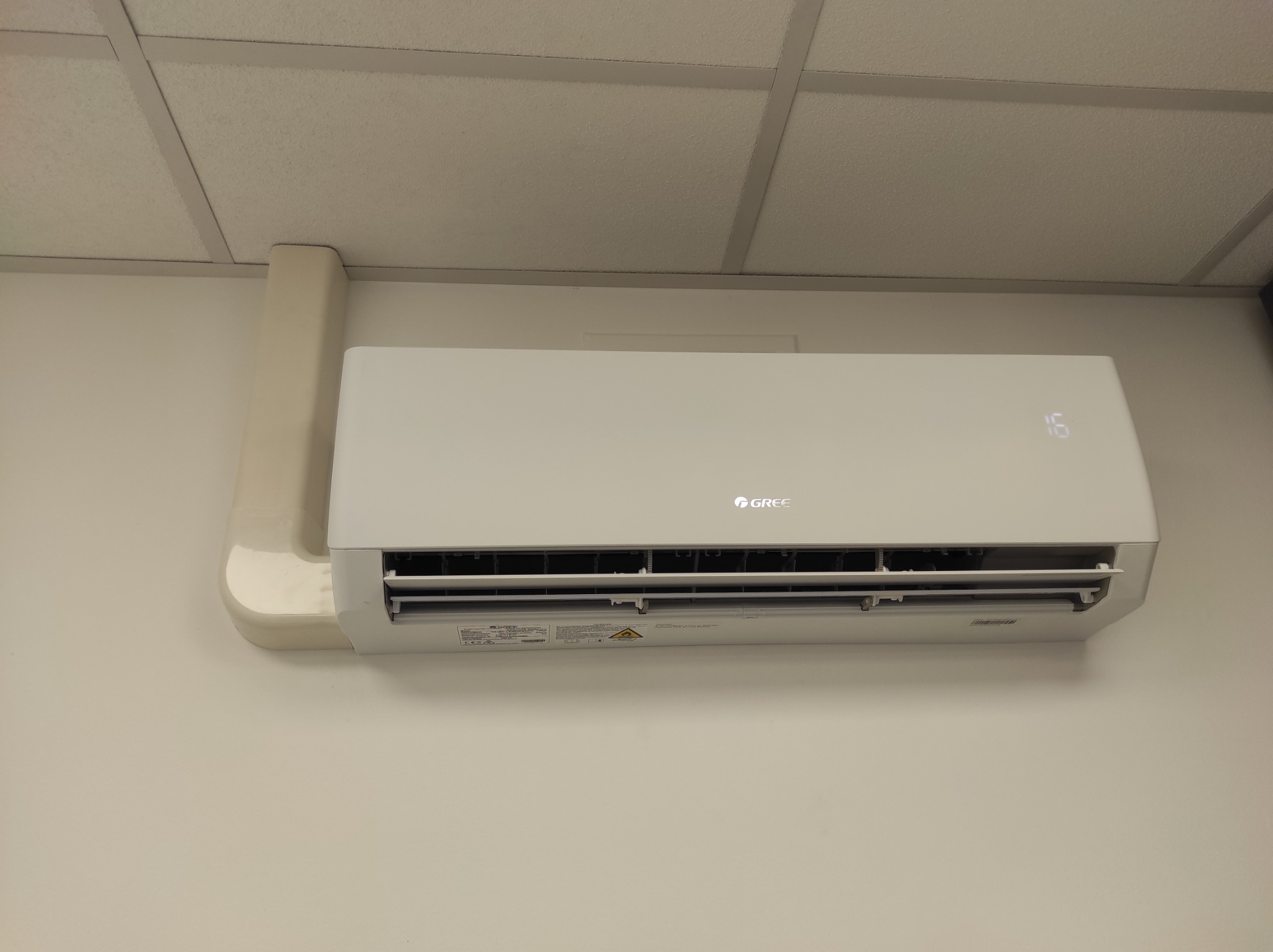Biała jednostka wewnętrzna klimatyzatora marki Gree zamontowana na jasnej ścianie, widoczny wyświetlacz z temperaturą 16 stopni Celsjusza i fragment instalacji odprowadzającej skropliny.