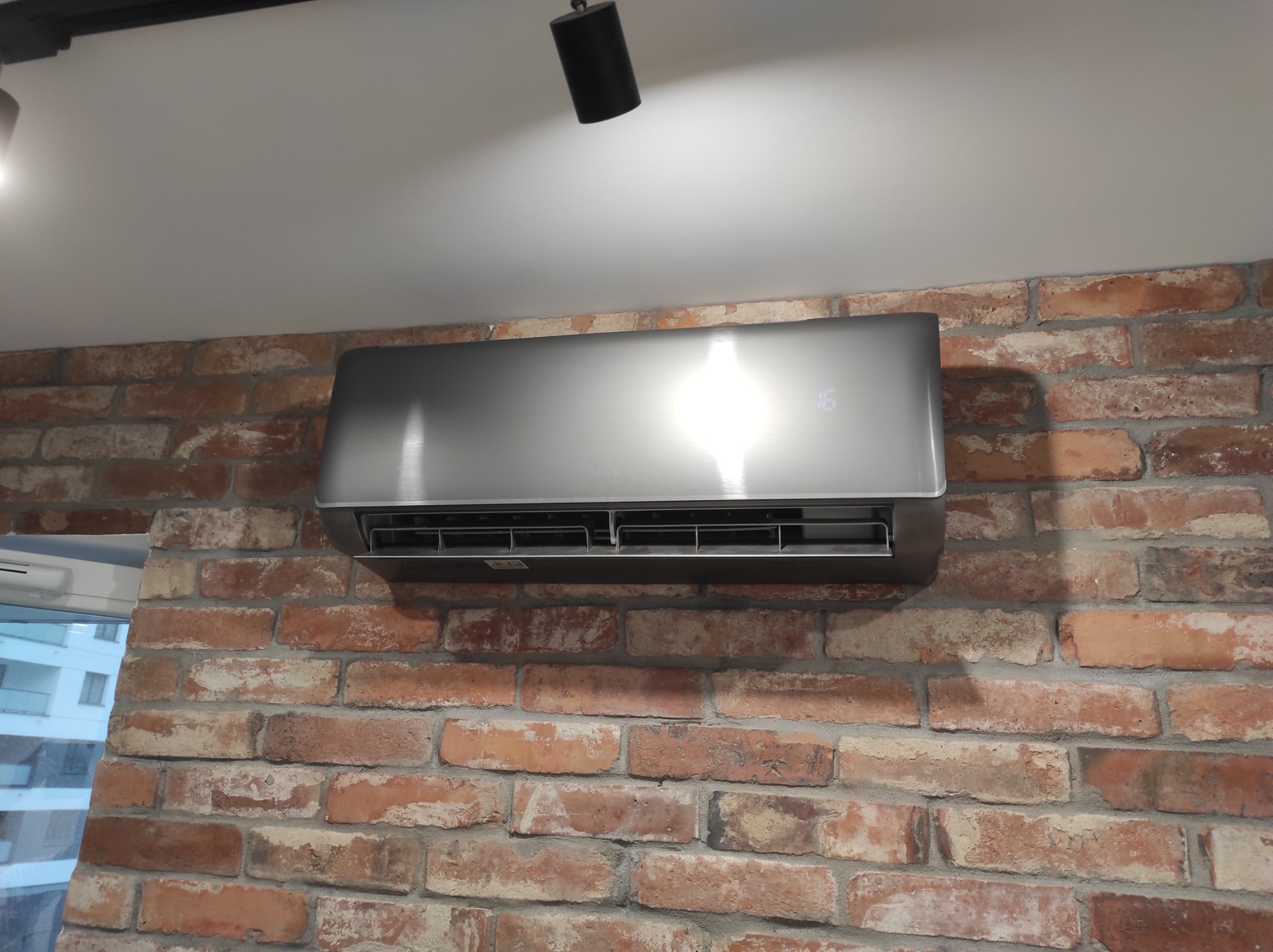 Szary klimatyzator ścienny z wyświetlaczem temperatury (15 stopni) zamontowany na ścianie z czerwonej cegły w pomieszczeniu z białym sufitem i oświetleniem punktowym.