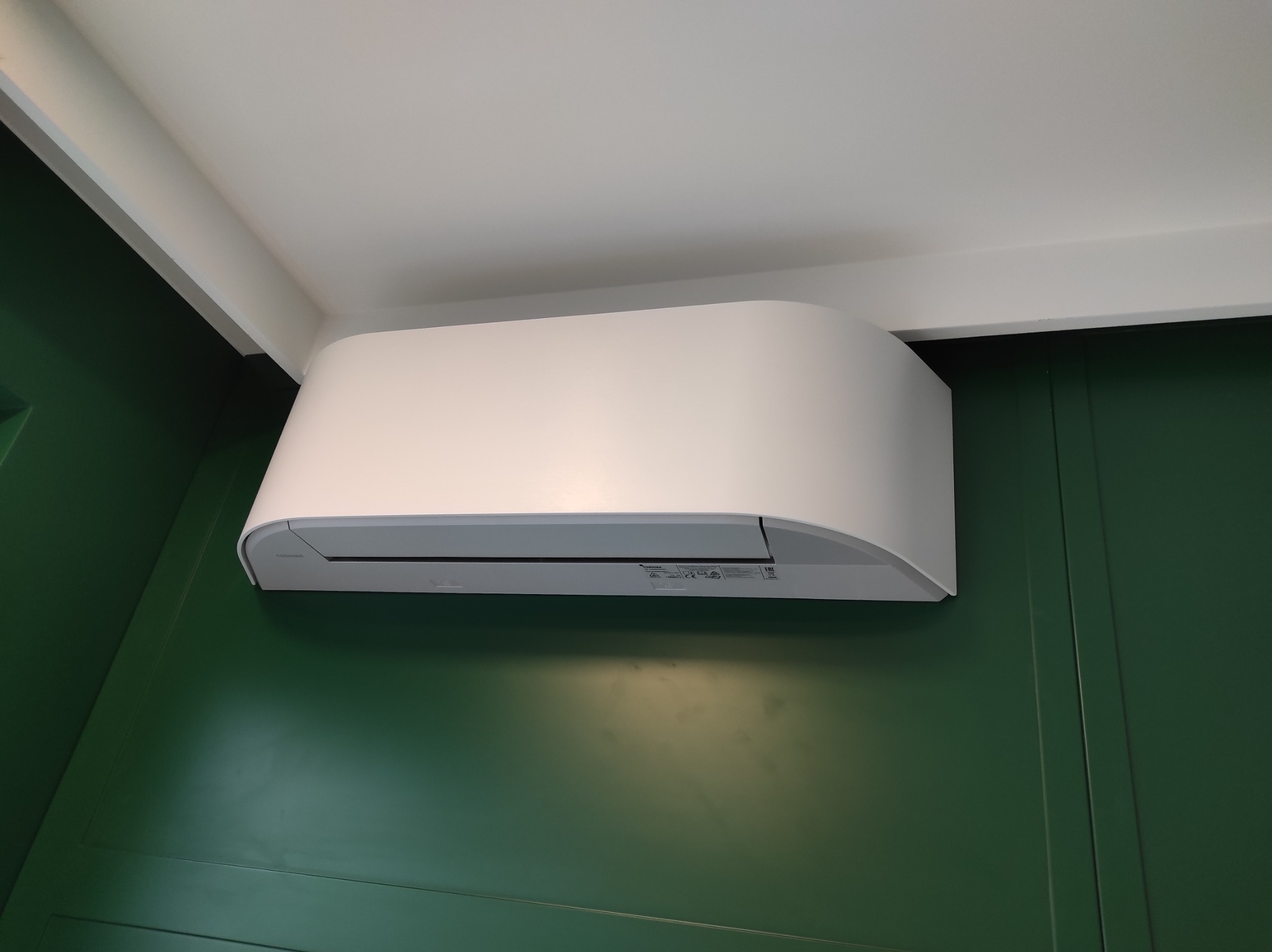 Biała jednostka wewnętrzna klimatyzacji zamontowana na ścianie w kolorze butelkowej zieleni z panelami dekoracyjnymi, widoczna spod sufitu.