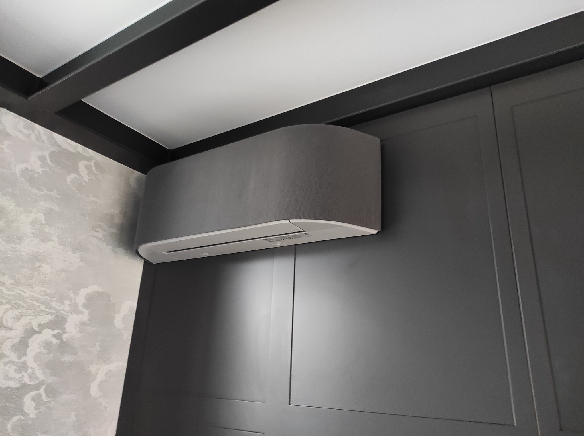 Szary klimatyzator ścienny zamontowany w rogu pokoju z dekoracyjną tapetą w chmury i ciemnymi listwami przysufitowymi.