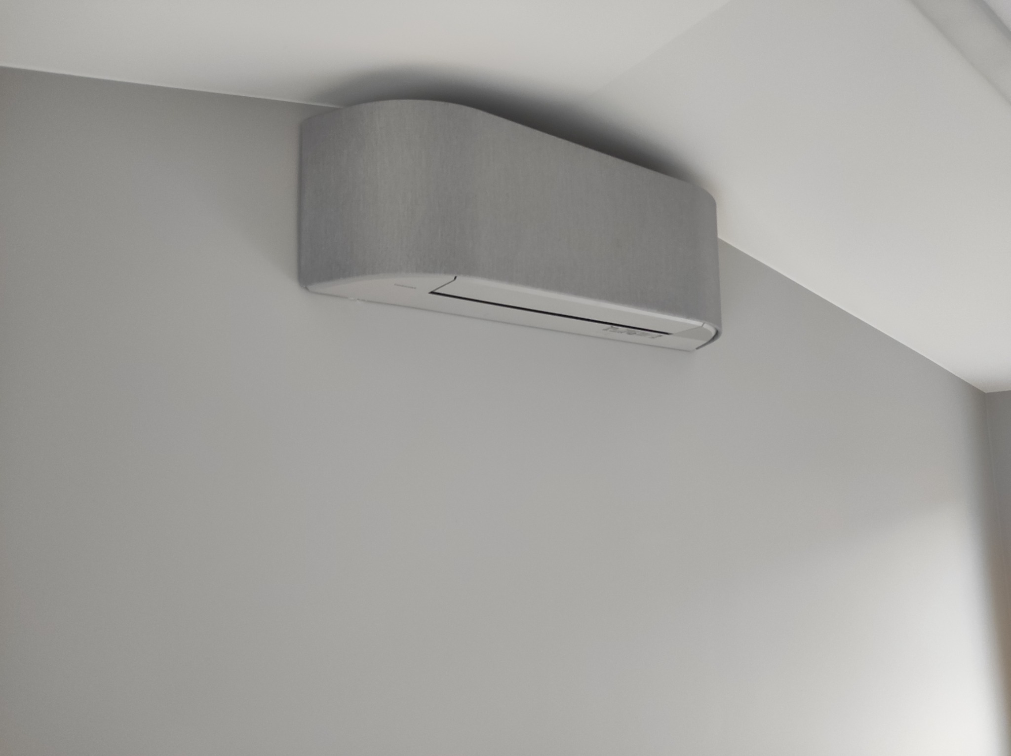 Szary klimatyzator ścienny o zaokrąglonych krawędziach, zamontowany w nowoczesnym wnętrzu na szarej ścianie, widok z dołu.