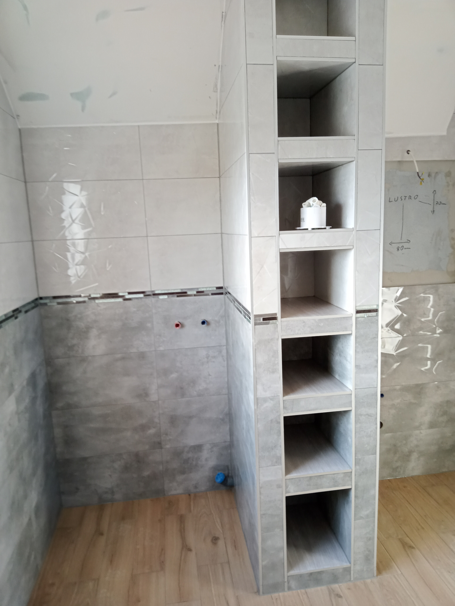 Wnętrze łazienki w trakcie remontu, widoczne płytki na ścianach w odcieniach szarości, drewnopodobna podłoga, wnęka z półkami obłożona płytkami oraz instalacja wodna. Na ścianie notatki dotyczące...