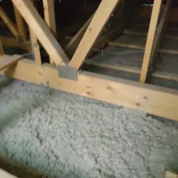 Wypełnienie przestrzeni między krokwiami izolacją wdmuchiwaną; widoczne drewniane elementy konstrukcji dachu i szara, włóknista izolacja pokrywająca podłogę poddasza.