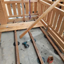 Budowa drewnianego tarasu: widok z góry na konstrukcję z legarów na regulowanych wspornikach, częściowo ułożone deski, wiertarka akumulatorowa i kątownik na betonowej posadzce.