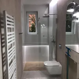 Nowoczesna łazienka z kabiną prysznicową typu walk-in, podświetlanym brodzikiem wyłożonym drewnem, wiszącą toaletą i grzejnikiem drabinkowym w kolorze białym. Okno z widokiem na drzewo na zewnątrz.