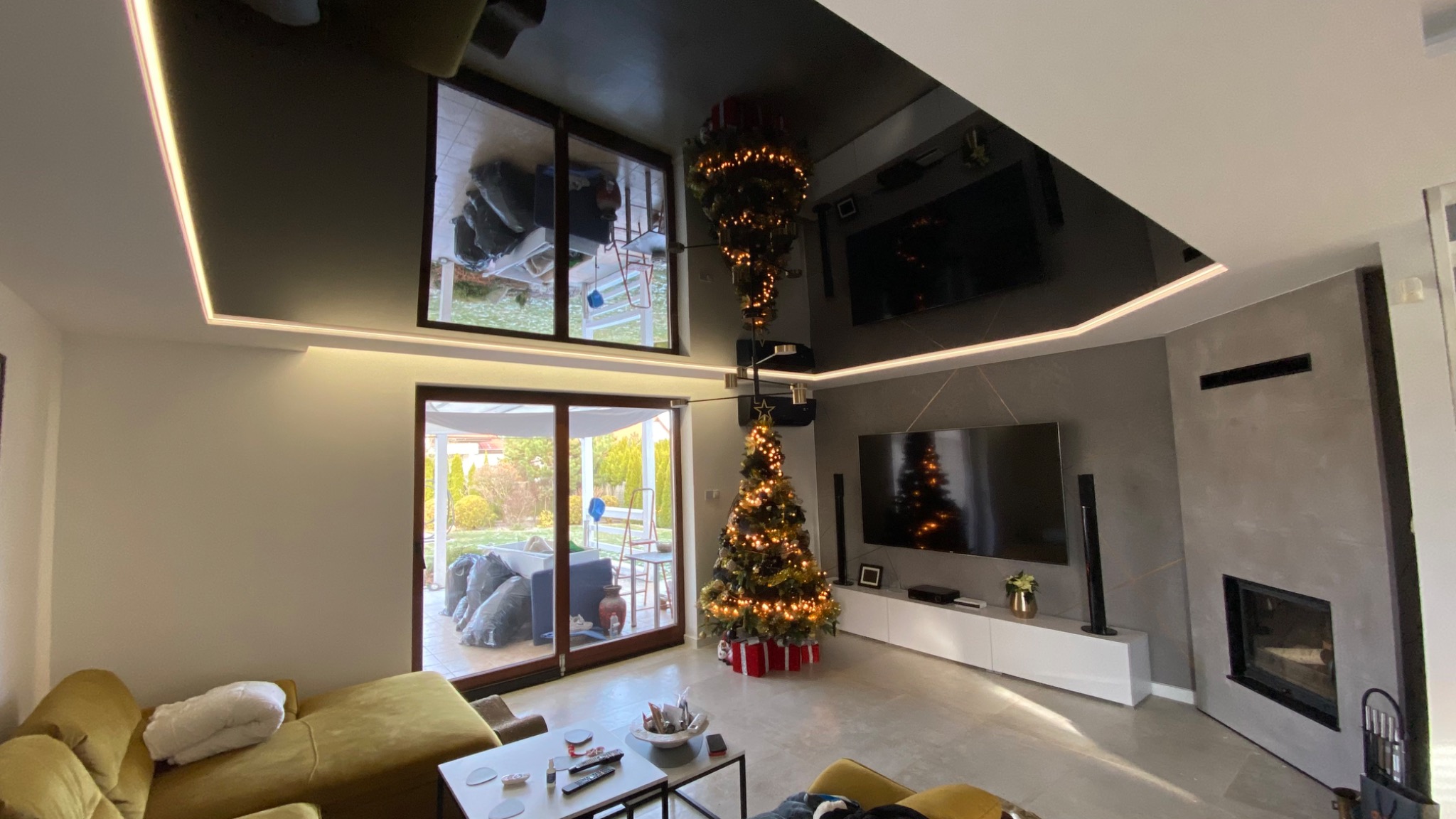 Nowoczesny salon z podwieszanym sufitem z oświetleniem LED, szarą ścianą z geometrycznym wzorem i kominkiem, oraz nietypową choinką zawieszoną do góry nogami obok tradycyjnej choinki.