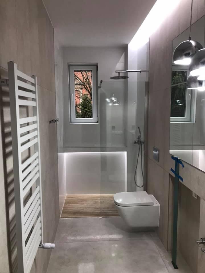 Nowoczesna łazienka z kabiną prysznicową typu walk-in, podświetlanym brodzikiem wyłożonym drewnem, wiszącą toaletą i grzejnikiem drabinkowym w kolorze białym. Okno z widokiem na drzewo na zewnątrz.