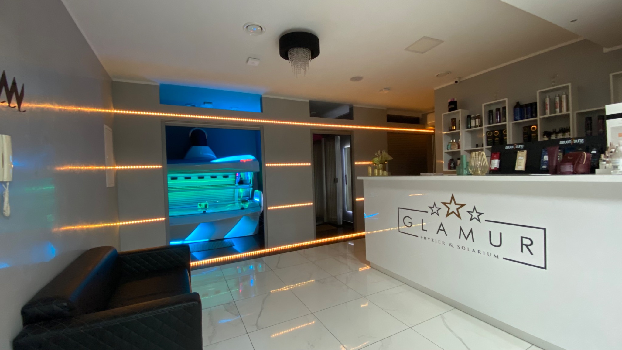 Wnętrze salonu fryzjersko-solaryjnego Glamur z recepcją, solarium z niebieskim światłem w tle, czarnym fotelem i oświetleniem LED.