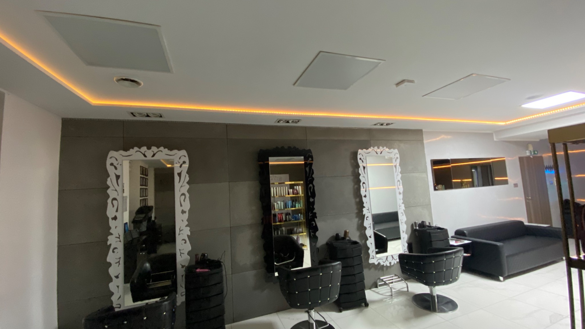 Wnętrze salonu fryzjerskiego z trzema stanowiskami, każde z ozdobnym lustrem i czarnym krzesłem na kółkach; sufit z podwieszanymi panelami i ledowym oświetleniem.