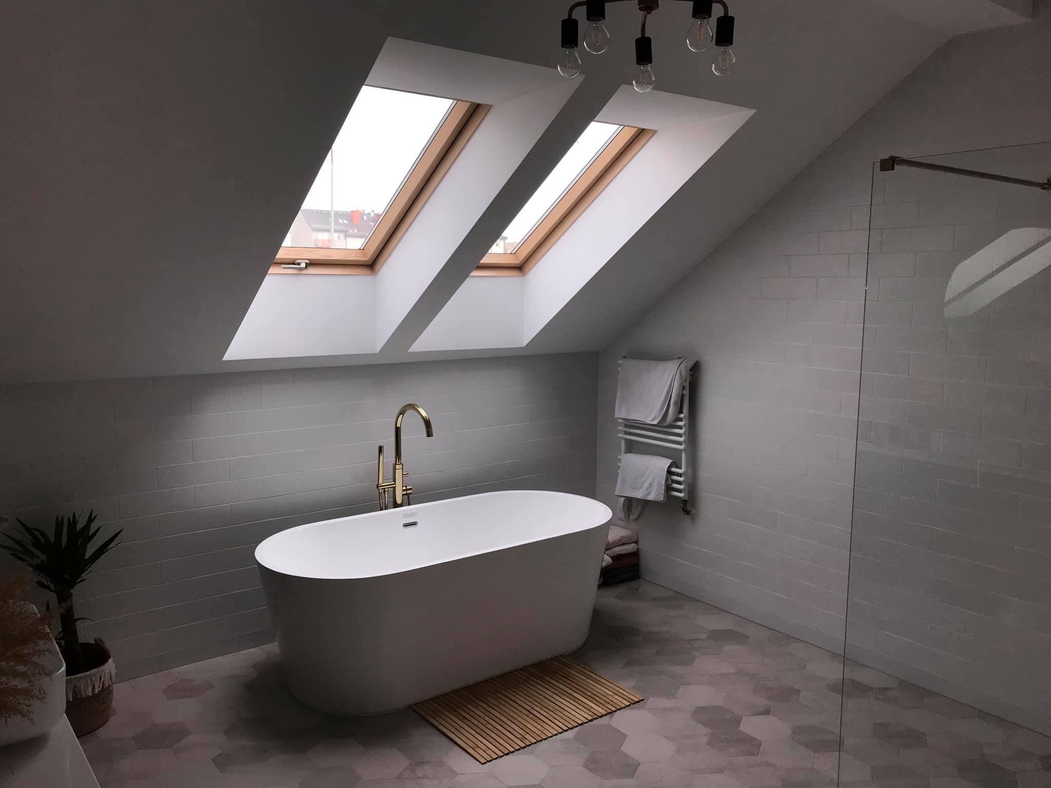 Nowoczesna łazienka na poddaszu z białą wanną wolnostojącą, złotą baterią i dwoma oknami dachowymi w drewnianych ramach. Na ścianach jasnoszare płytki imitujące cegłę, na podłodze heksagonalne...