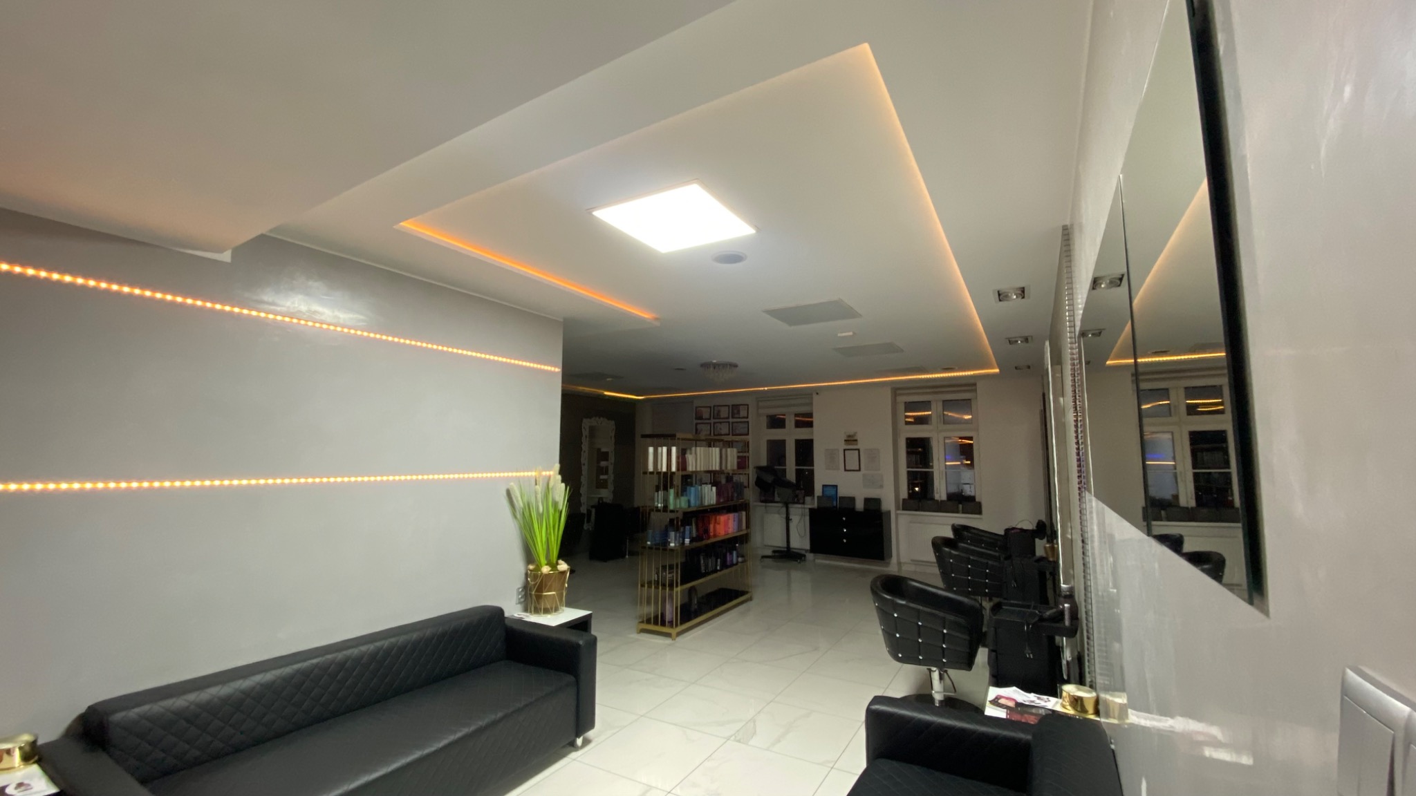 Nowoczesne wnętrze salonu fryzjerskiego z szarą ścianą, podświetlanymi ledami, czarną sofą z pikowaniem, regałem z kosmetykami i lustrami.