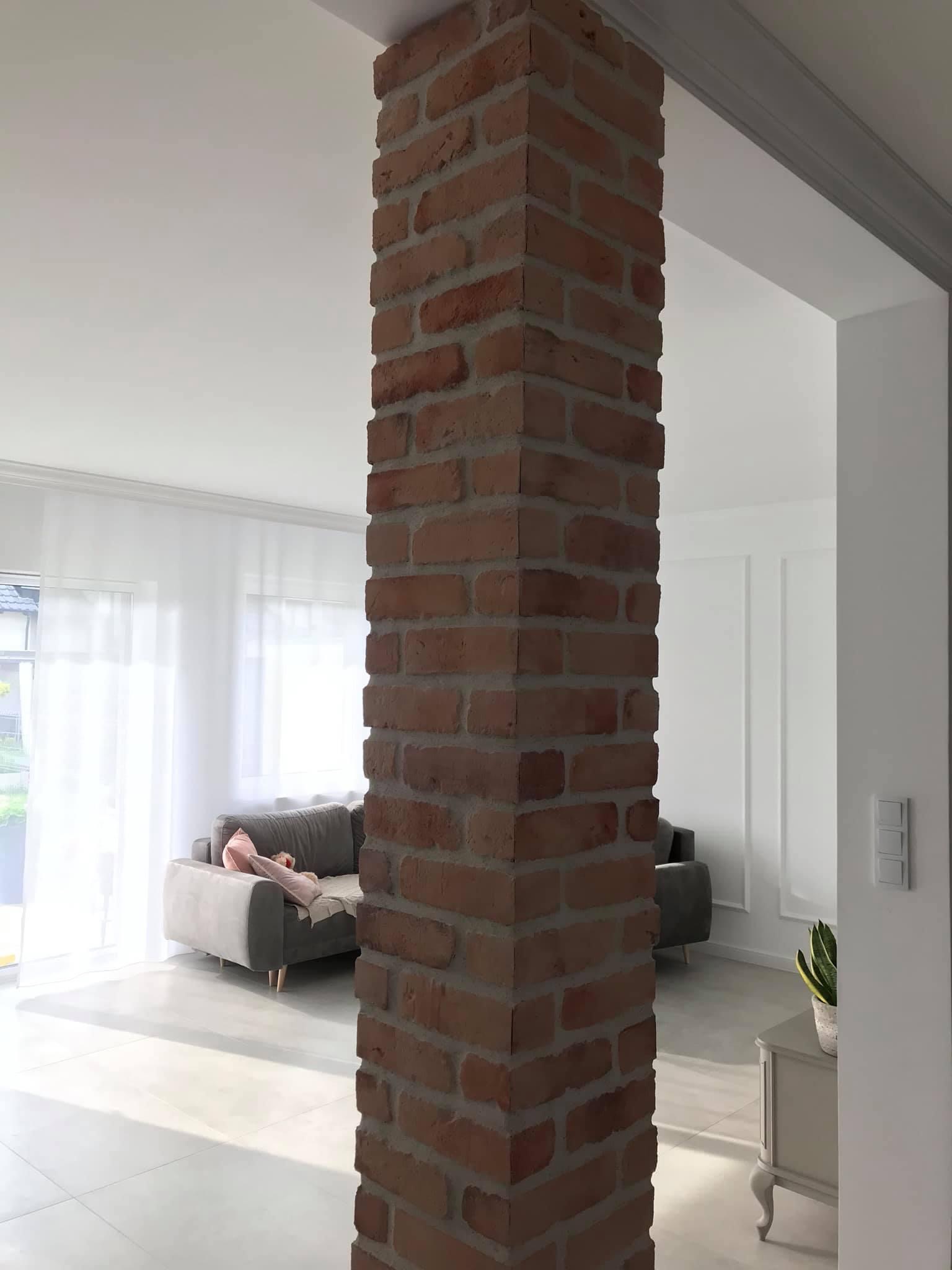 Wnętrze salonu z ceglaną kolumną, widoczna sofa, firanki i białe ściany z dekoracyjnymi listwami.