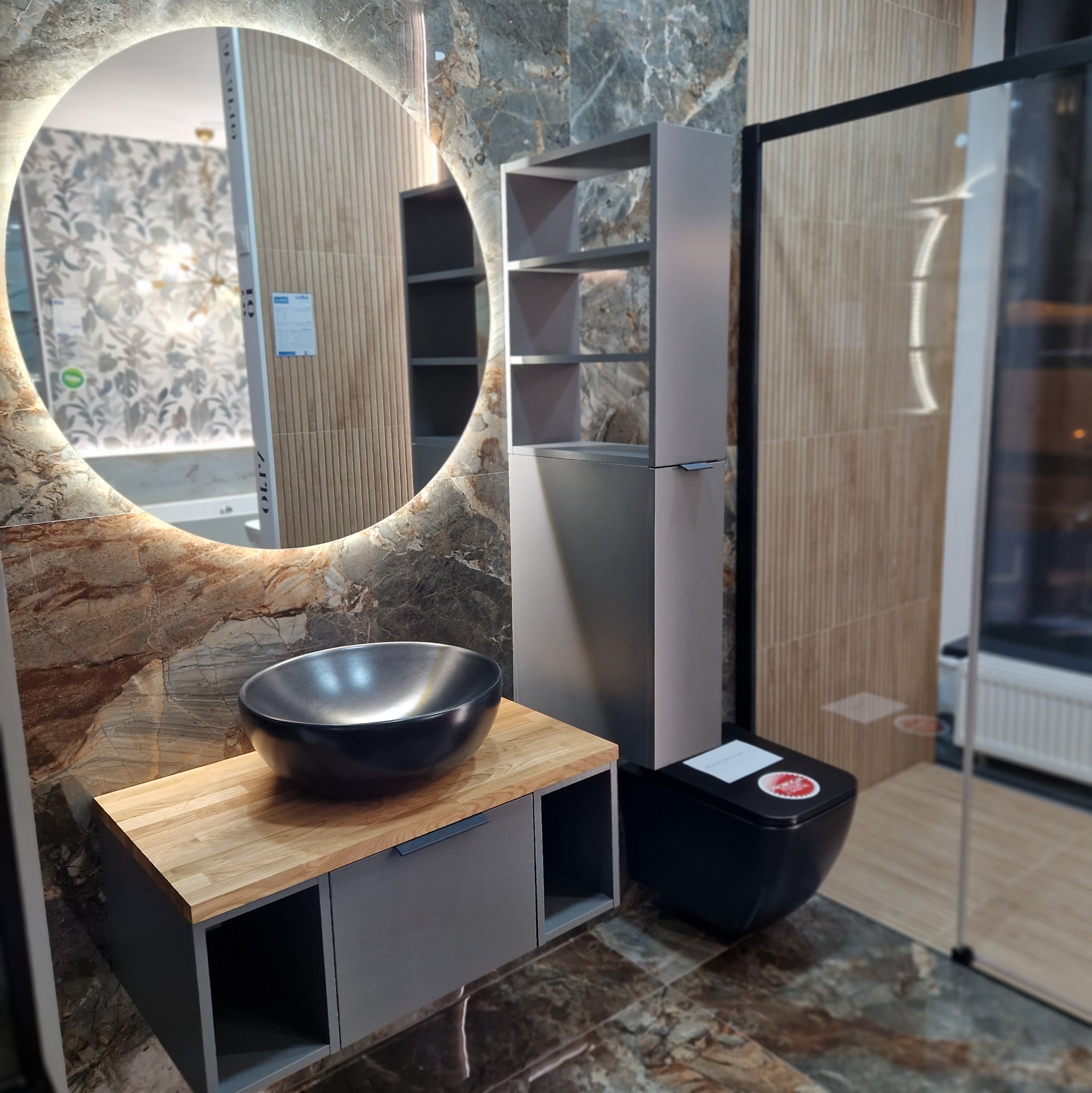 Nowoczesna łazienka z okrągłym lustrem podświetlanym LED, umywalką w formie misy, szafką z drewnianym blatem i czarną toaletą.