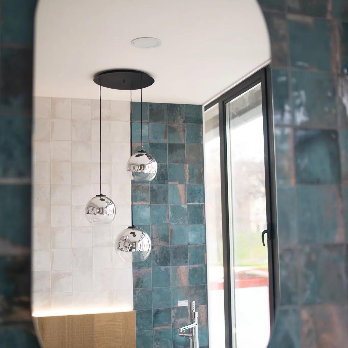 Nowoczesna łazienka z turkusowymi i białymi kwadratowymi płytkami, wisząca lampa z trzema szklanymi kulami, widoczna w lustrze.