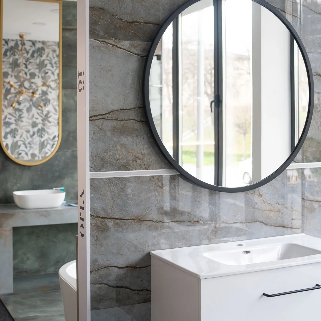 Nowoczesna łazienka z okrągłym lustrem w czarnej ramie, białą szafką pod umywalką i płytkami imitującymi marmur z widoczną armaturą.