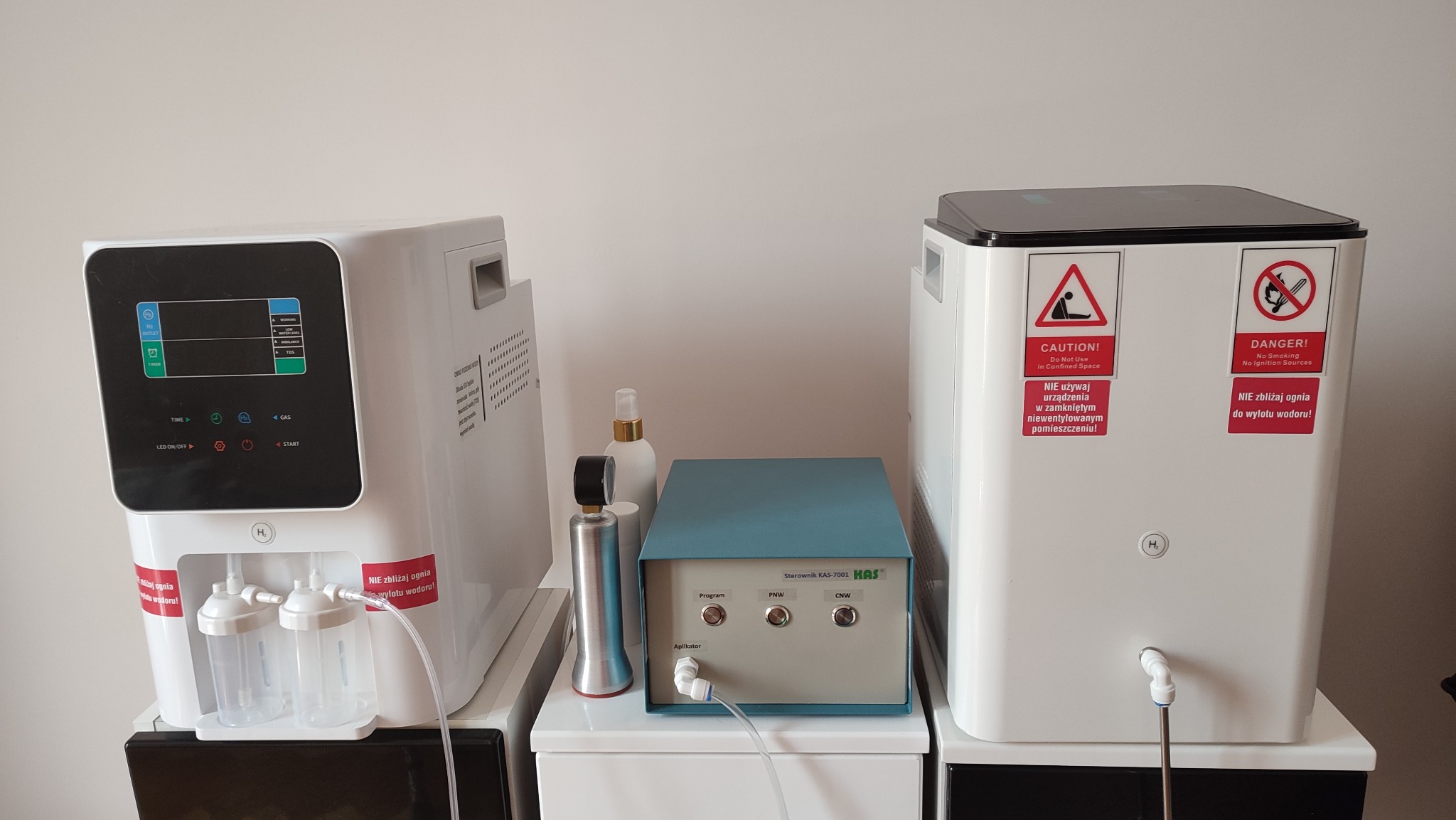 Dwa białe generatory wodoru, jeden z wyświetlaczem dotykowym i pojemnikami na wodę, drugi z naklejkami ostrzegawczymi, oraz niebieski sterownik KAS-7001 HAS z przyciskami i aplikatorem.