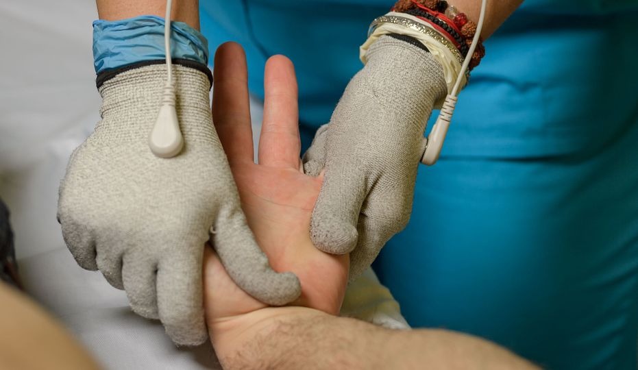 Terapia dłoni przy użyciu elektrostymulacji; rękawice przewodzące z elektrodami na dłoni pacjenta, niebieskie ubranie terapeuty i bransoletki na nadgarstku.