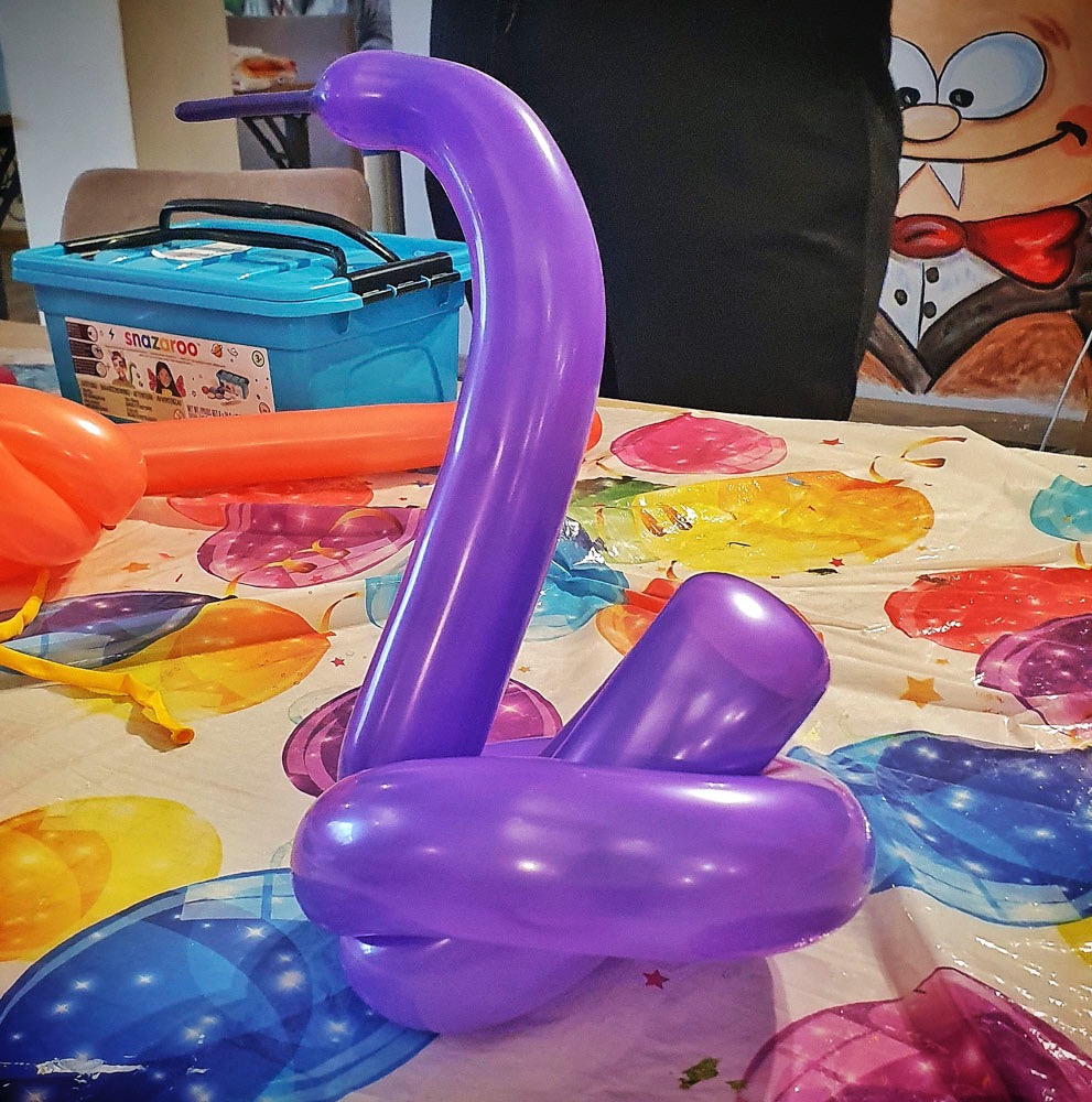 Fioletowy balon uformowany w kształt łabędzia, ustawiony na stole z kolorowym obrusem, obok pudełka z akcesoriami do modelowania balonów.