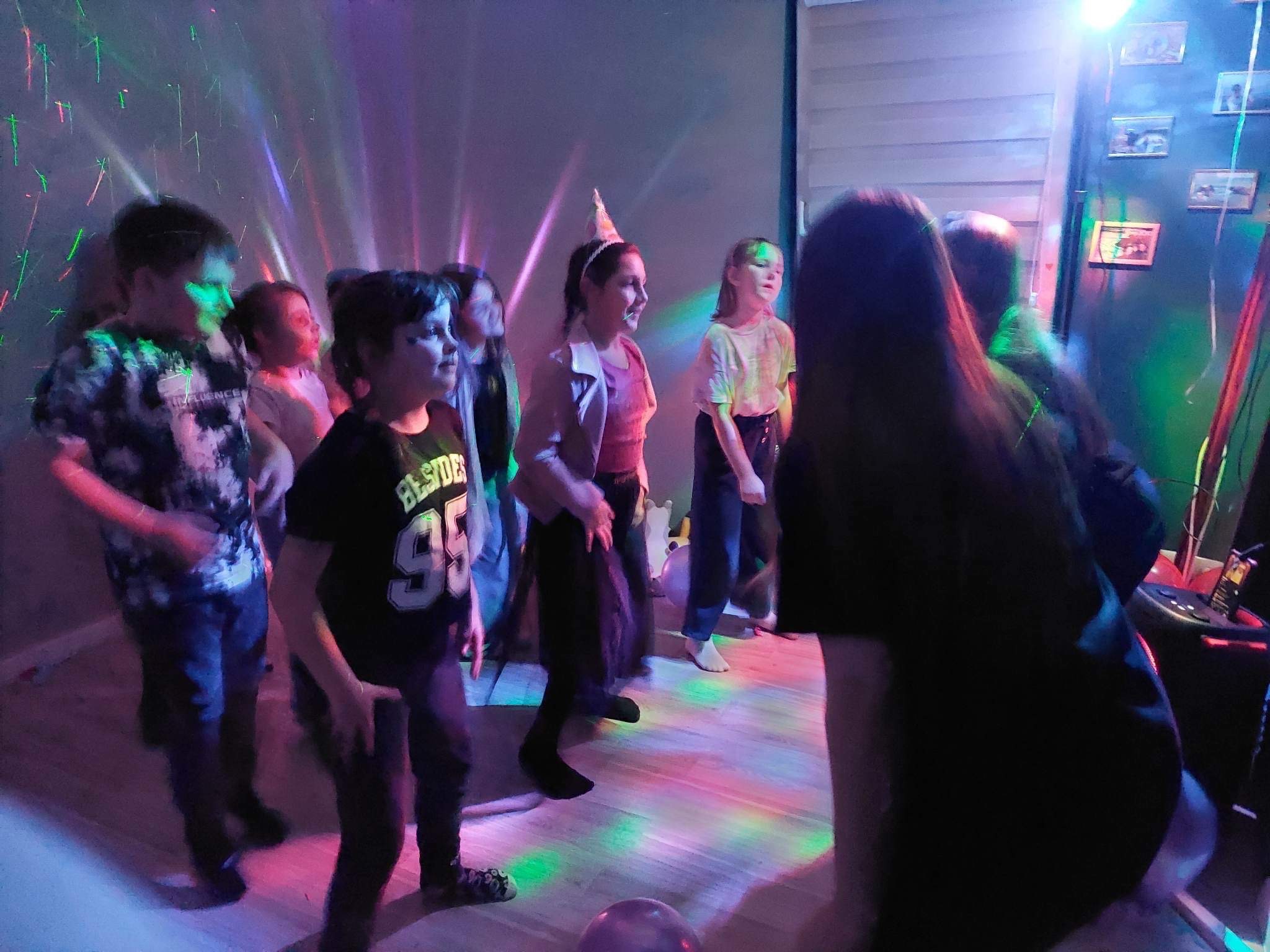 Grupa dzieci tańczy w pomieszczeniu oświetlonym kolorowymi światłami dyskotekowymi, jedno dziecko ma na głowie papierową czapeczkę urodzinową, na ścianie wiszą zdjęcia.