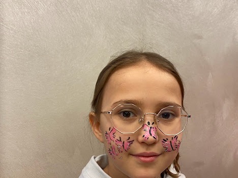 Dziewczynka w okularach z pomalowaną twarzą w różowe motyle i czarne kontury, po wykonaniu usługi malowania twarzy.