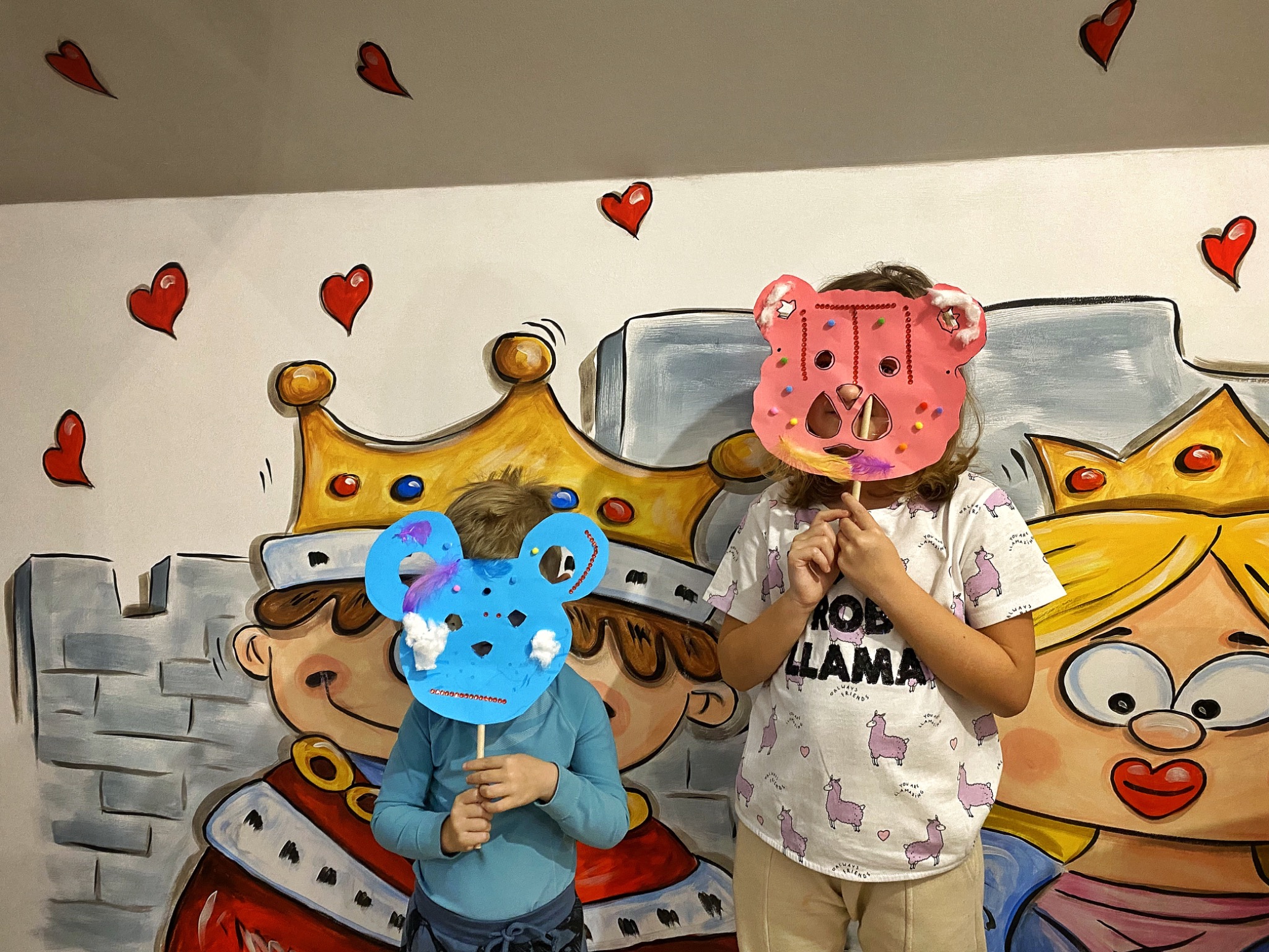 Dwoje dzieci trzymających przed twarzami wykonane własnoręcznie maski na patykach, stojących na tle ściany z namalowaną parą królewską i czerwonymi sercami.