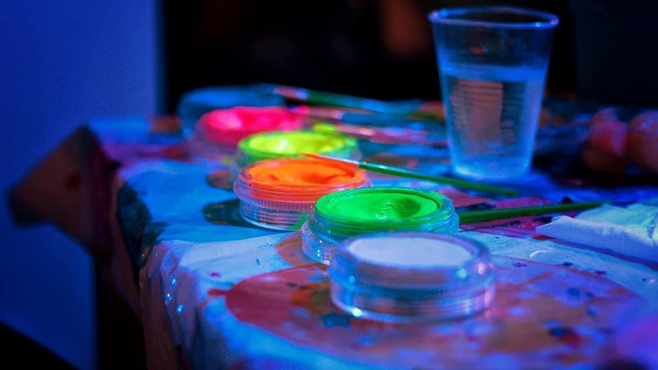 Przygotowane farby do malowania twarzy w neonowych kolorach w plastikowych pojemnikach na stole, obok pędzle i szklanka wody.