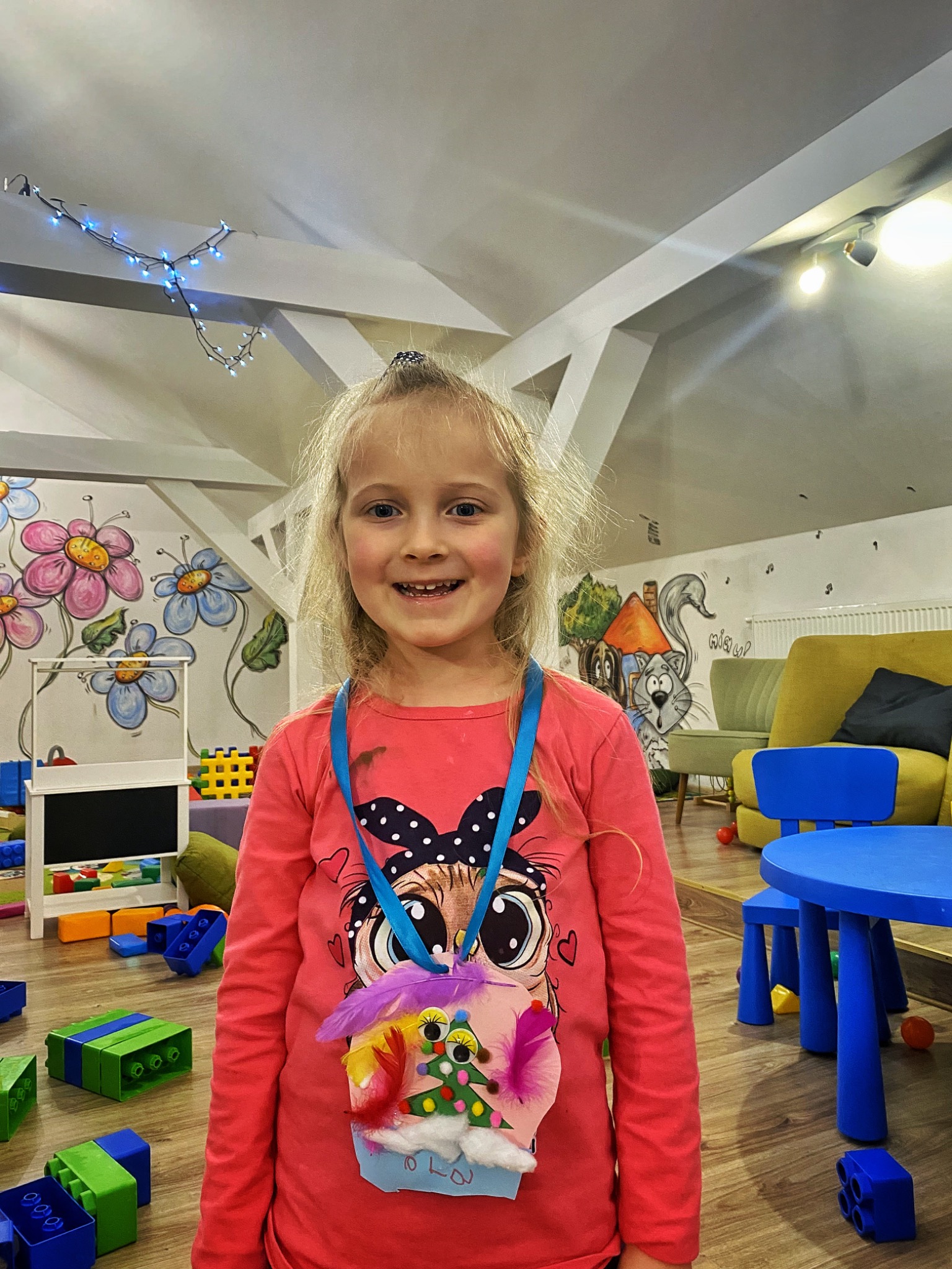 Uśmiechnięta dziewczynka z blond włosami, w różowej bluzce z aplikacją, prezentuje własnoręcznie zrobiony naszyjnik z piórek i choinką, w tle pokój zabaw z klockami i malowidłami na ścianach.