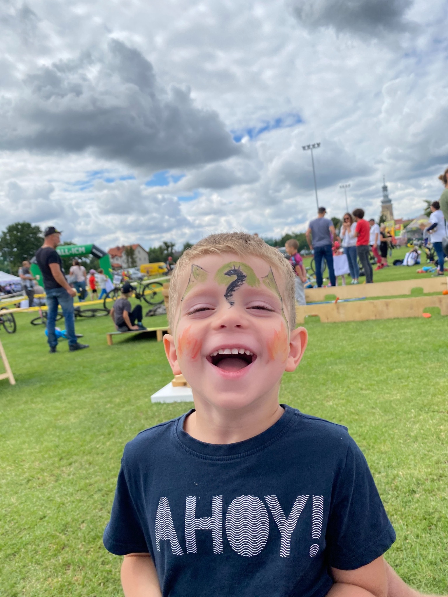 Rozesmiany chłopiec z pomalowaną twarzą w smoka i pomarańczowe plamy na policzkach, na tle zielonego trawnika, ludzi i kościoła w oddali.