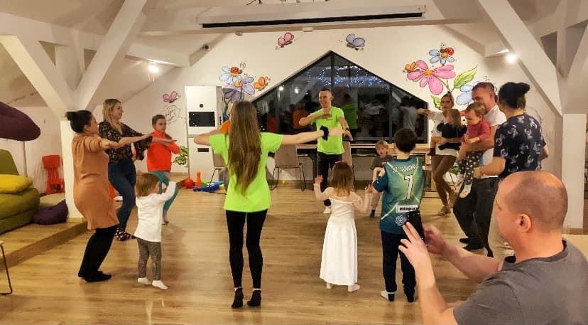 Zabawa taneczna z udziałem dzieci i dorosłych w sali z dekoracjami kwiatowymi i motylami na ścianie w Sosnówce. Osoba prowadząca w neonowej koszulce pokazuje ruchy.