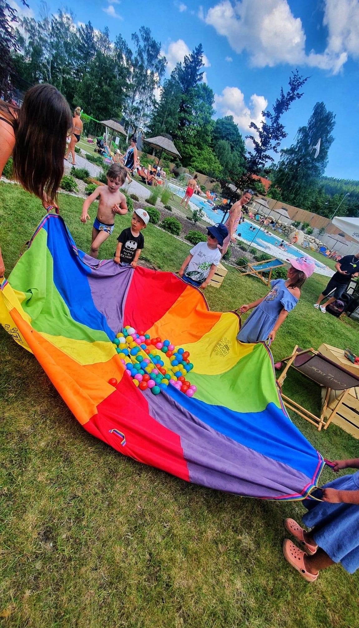 Zabawa z chustą animacyjną w plenerze: grupa dzieci trzyma kolorową chustę z tęczowymi segmentami, na której leżą kolorowe piłeczki; w tle basen i wypoczywający ludzie.