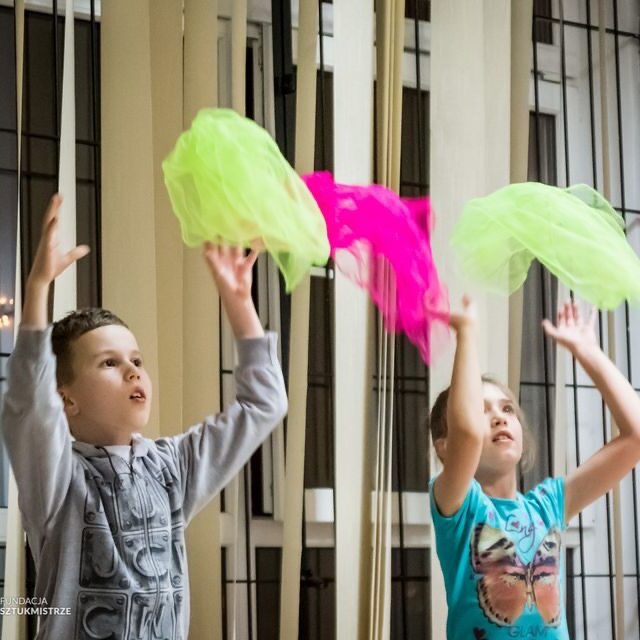 Dwoje dzieci, chłopiec w szarej bluzie i dziewczynka w niebieskiej koszulce z motylem, uczą się żonglować kolorowymi chustami w jasnym pomieszczeniu z oknami.