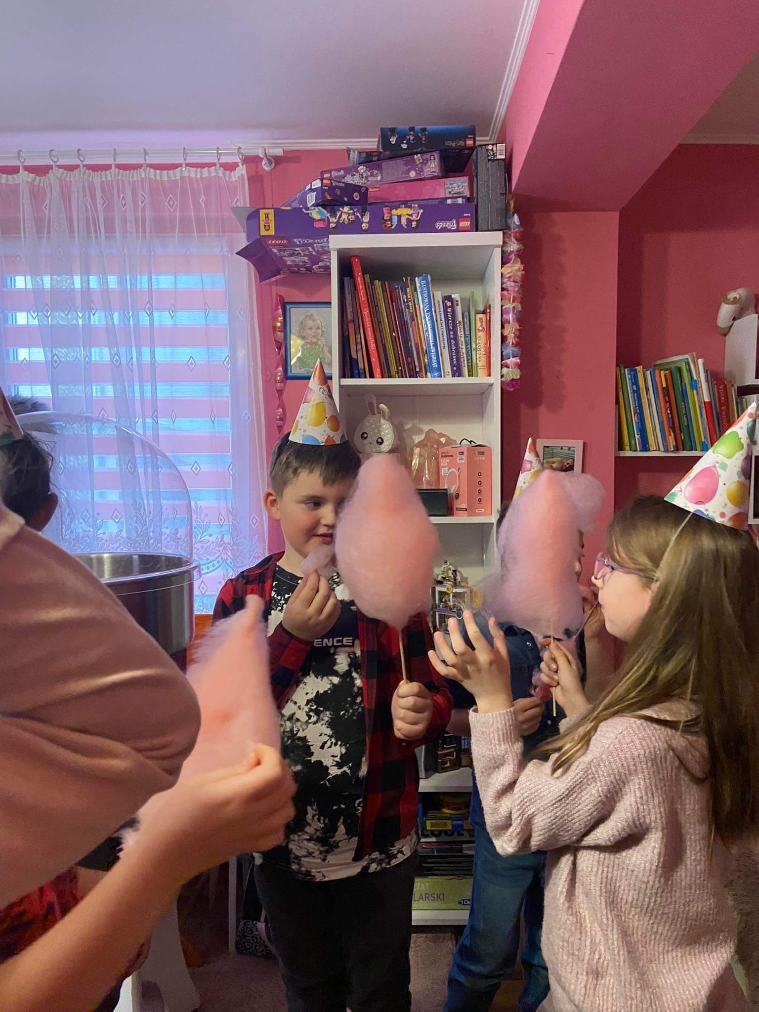 Dzieci w papierowych czapeczkach zajadają watę cukrową podczas urodzinowego przyjęcia w pokoju z różowymi ścianami i regałem pełnym książek i pudełek Lego Friends.