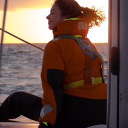 Naturalnie Sentymentalnie Natalia Sajkowska Fotografia - Kobieta w pomarańczowej kurtce i uprzęży siedzi na jachcie, z zamkniętymi oczami i uśmiechem, w tle zachodzące słońce nad morzem.