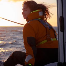 Naturalnie Sentymentalnie Natalia Sajkowska Fotografia - Kobieta w pomarańczowej kurtce i uprzęży siedzi na jachcie, patrząc w dal na zachodzące słońce nad morzem.