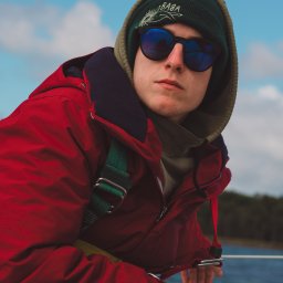 Naturalnie Sentymentalnie Natalia Sajkowska Fotografia - Młody mężczyzna w czerwonej kurtce, zielonej czapce i okularach przeciwsłonecznych spogląda w obiektyw na tle błękitnego nieba i wody, trzymając w ręku biały przedmiot.