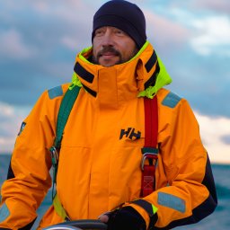 Naturalnie Sentymentalnie Natalia Sajkowska Fotografia - Mężczyzna w pomarańczowej kurtce żeglarskiej i czarnej czapce trzyma ster jachtu na tle pochmurnego nieba i wody.