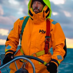 Naturalnie Sentymentalnie Natalia Sajkowska Fotografia - Mężczyzna w pomarańczowym sztormiaku marki HH, z papierosem w ustach, steruje jachtem na tle zachmurzonego nieba o zmierzchu.