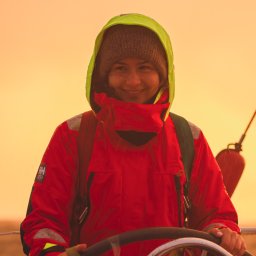 Naturalnie Sentymentalnie Natalia Sajkowska Fotografia - Uśmiechnięta kobieta w czerwonej kurtce żeglarskiej i wełnianej czapce, trzymająca ster jachtu podczas rejsu o zachodzie słońca w okolicach Lubania.
