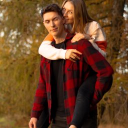 Naturalnie Sentymentalnie Natalia Sajkowska Fotografia - Uśmiechnięta kobieta całuje w policzek mężczyznę niosącego ją na barana, jesienne tło z drzewami w odcieniach złota i brązu, widoczny tatuaż na kostce mężczyzny.