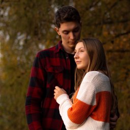 Naturalnie Sentymentalnie Natalia Sajkowska Fotografia - Młoda para w jesiennej scenerii: mężczyzna w czerwonej koszuli w kratę obejmuje kobietę w swetrze w biało-pomarańczowe pasy, oboje zamyśleni.