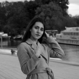 Naturalnie Sentymentalnie Natalia Sajkowska Fotografia - Portret kobiety w płaszczu na tle nabrzeża i statku, zdjęcie czarno-białe, z delikatnym uśmiechem i ręką przy włosach.