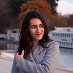 Naturalnie Sentymentalnie Natalia Sajkowska Fotografia - Portret uśmiechniętej kobiety w szarym płaszczu na tle jesiennego parku i rzeki z łodziami.