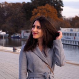 Naturalnie Sentymentalnie Natalia Sajkowska Fotografia - Uśmiechnięta kobieta w szarym płaszczu, z torebką na łańcuszku, poprawia włosy na tle jesiennego krajobrazu z wodą i budynkami.