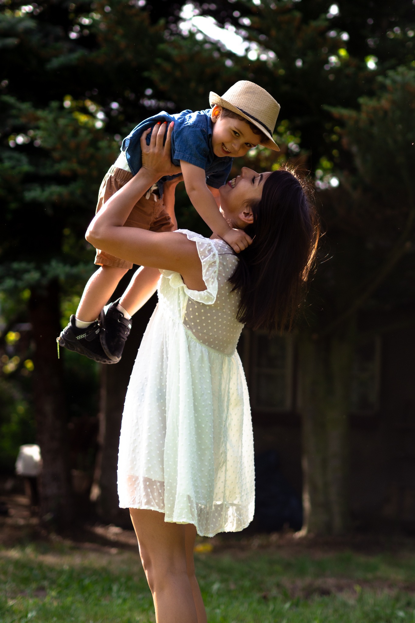 Uśmiechnięta matka w białej sukience podnosi do góry synka w słomkowym kapeluszu na tle zieleni.