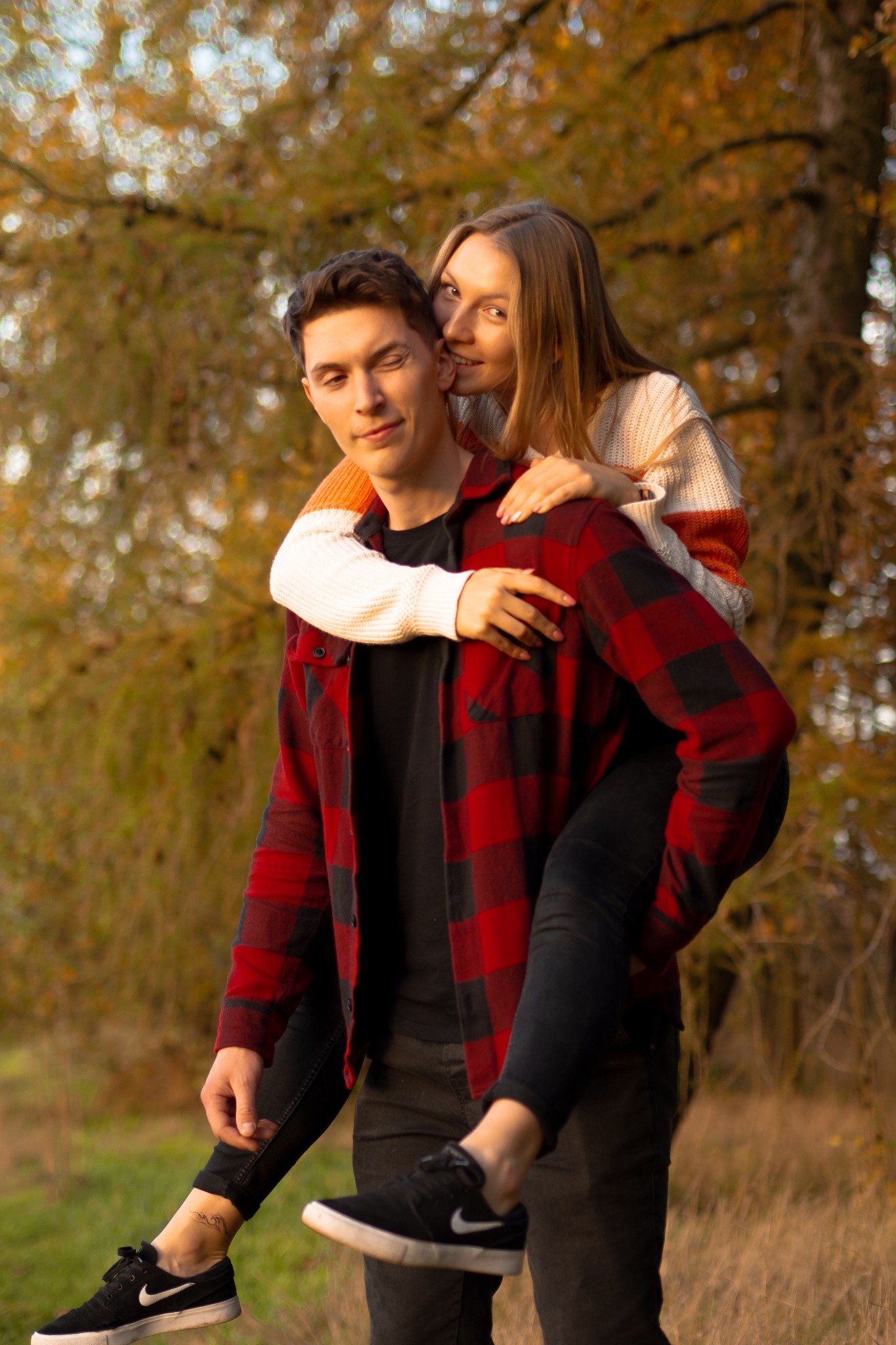 Uśmiechnięta kobieta całuje w policzek mężczyznę niosącego ją na barana, jesienne tło z drzewami w odcieniach złota i brązu, widoczny tatuaż na kostce mężczyzny.
