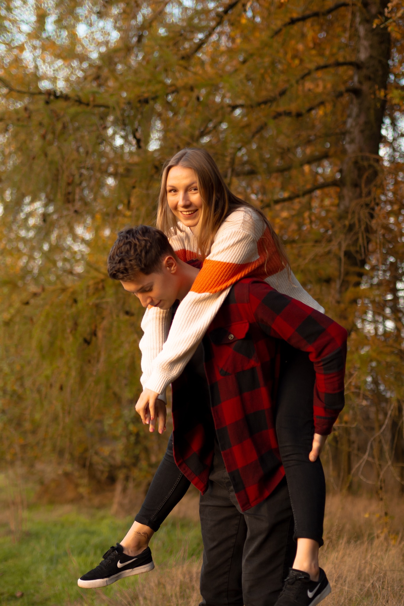 Uśmiechnięta blondynka na barana u młodego mężczyzny w parku, jesienne drzewa w tle, oboje w casualowych ubraniach, ona w kremowym swetrze w pomarańczowe pasy, on w czerwonej koszuli w kratę.