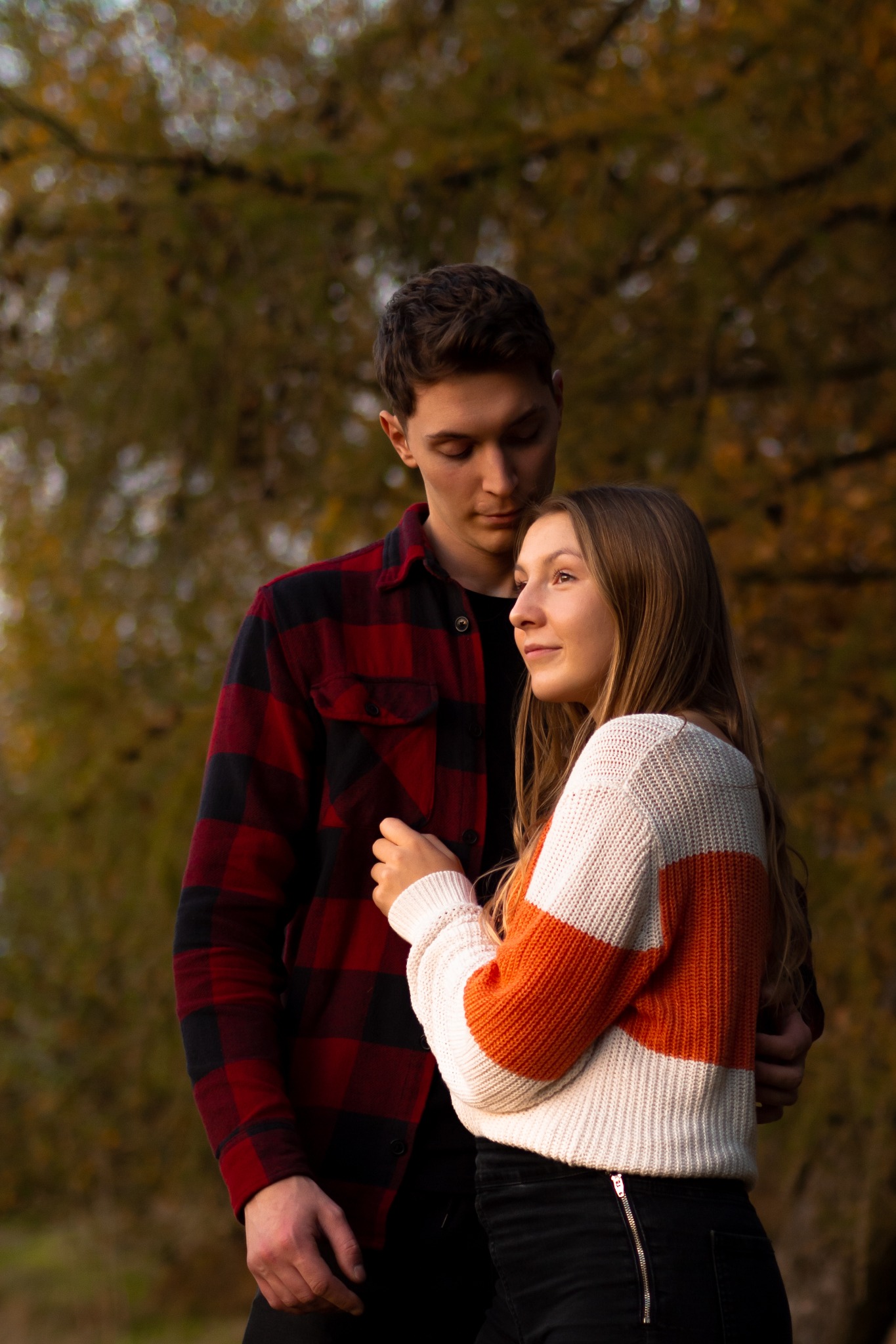 Młoda para w jesiennej scenerii: mężczyzna w czerwonej koszuli w kratę obejmuje kobietę w swetrze w biało-pomarańczowe pasy, oboje zamyśleni.