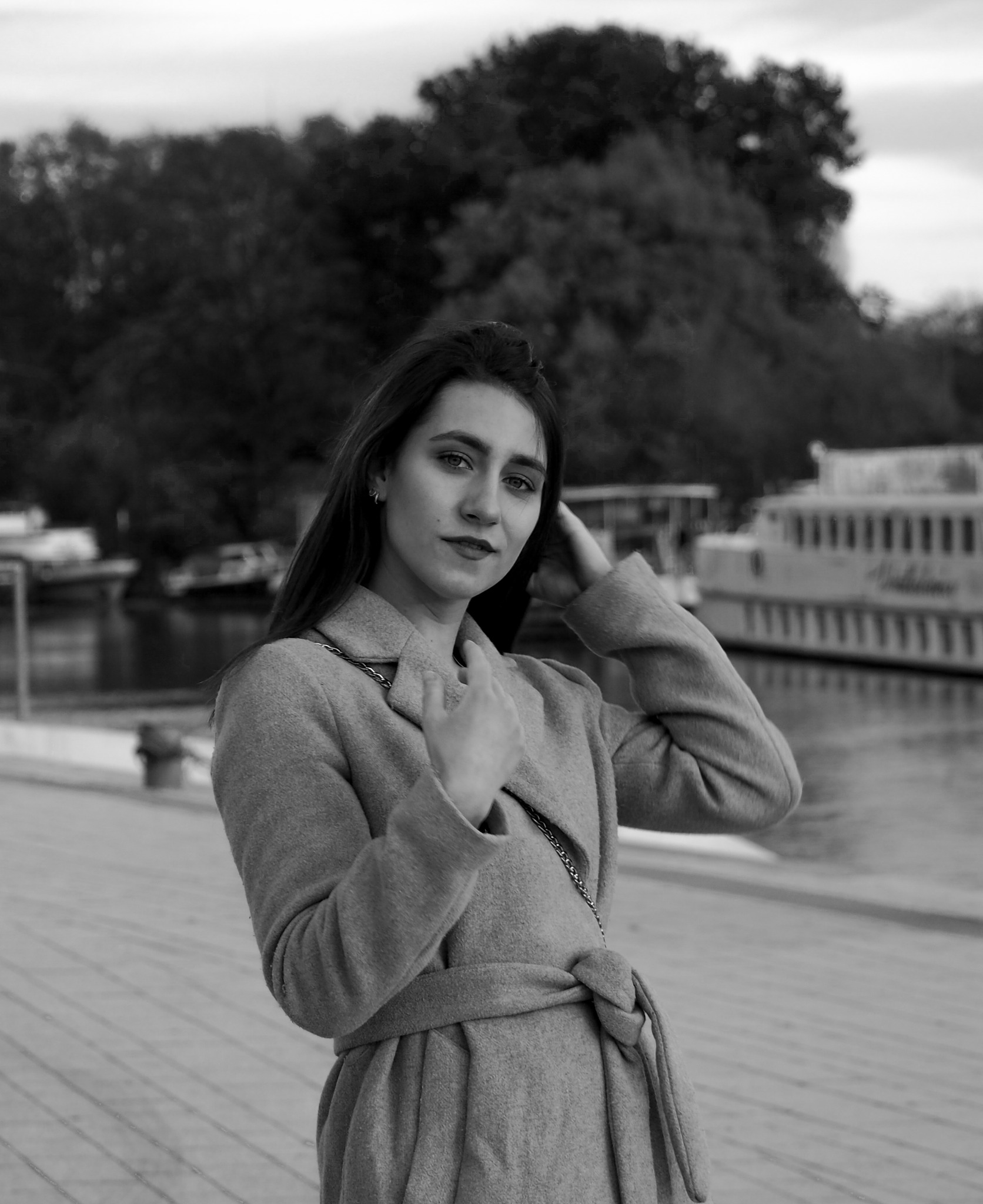 Portret kobiety w płaszczu na tle nabrzeża i statku, zdjęcie czarno-białe, z delikatnym uśmiechem i ręką przy włosach.